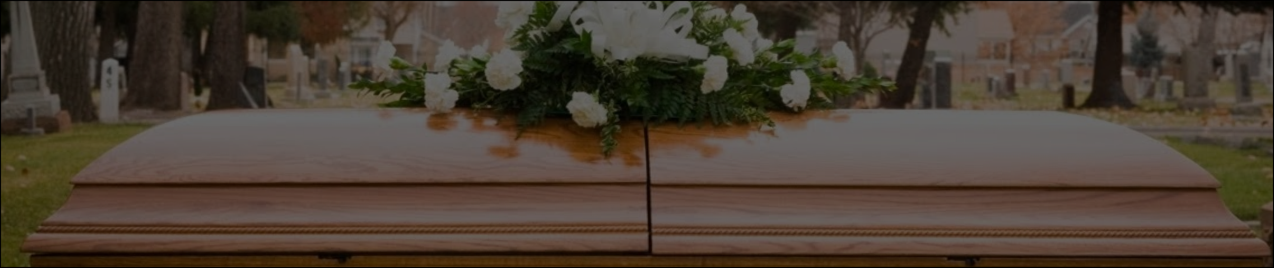 Coffin with white flowers on top in a cemetery with trees and gravestones in the background